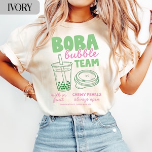 May include: Ivory-colored t-shirt with a green and pink graphic that says "BOBA bubble TEAM". The graphic includes a boba drink and a container with the text "milk or fruit" and "CHEWY PEARLS always open".