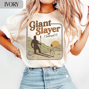 Op de afbeelding: Ivoor T-shirt met een grafisch ontwerp. Het ontwerp toont de woorden "Giant Slayer" en "1 Samuel 17". Het toont een reus en een kleinere figuur in een woestijnlandschap met een ondergaande zon.