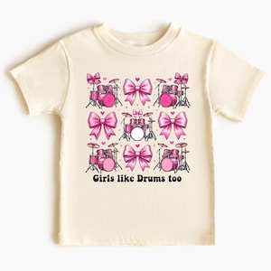 Könnte beinhalten: Cremefarbenes T-Shirt mit einem sich wiederholenden Muster aus rosa Schlagzeugsets und großen rosa Schleifen. Der Text "Girls like Drums too" ist darunter in Schwarz gedruckt. Das Shirt besteht aus einem weichen Material.