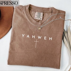 Pode incluir: Camiseta cor de café com a palavra "YAHWEH" e uma cruz impressa em branco. A camiseta tem gola redonda e mangas curtas. Um colar de prata com um pingente está drapeado sobre a camiseta.