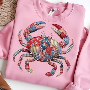 May include: Pink sweatshirt featuring a patchwork crab design. The crab is made of various fabric patterns in red, blue, and yellow. The sweatshirt has a crew neck and long sleeves.