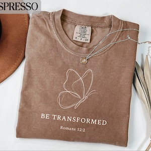 May include: Espresso-colored t-shirt with a white butterfly outline and the words "BE TRANSFORMED" and "Romans 12:2". The shirt has a crew neck and a small tag. A silver necklace with a pendant is on the shirt.