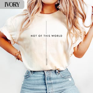 Op de afbeelding: Ivoor kleurig T-shirt met de zwarte tekst "NOT OF THIS WORLD" gecentreerd. Een verticale lijn loopt van de tekst, met "JOHN 18:36" eronder gedrukt. Het shirt heeft korte mouwen en wordt gecombineerd met een blauwe jeans.