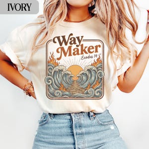 Pode incluir: Camiseta de cor marfim com um design gráfico castanho com as palavras "Way Maker" e "Exodus 14". O design inclui um sol, ondas e chamas. A camiseta é combinada com jeans azul.