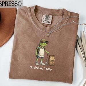 Puede incluir: Camiseta color espresso con un diseño de rana con sombrero y delantal, sosteniendo una espátula. La frase "No Grilling Today" está impresa debajo. Un collar plateado está sobre la camiseta.