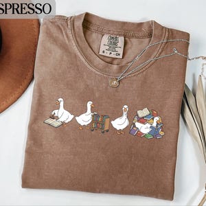 May include: Espresso-coloured t-shirt with a crew neck and short sleeves. The shirt features a graphic of three white geese interacting with books. One goose reads a book, another pushes a cart of books, and the third is surrounded by books.