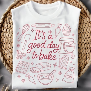 Könnte beinhalten: Weißes T-Shirt mit einer roten Strichzeichnung von Backutensilien und dem Schriftzug "It's a good day to bake". Das Design umfasst einen Schneebesen, eine Teigrolle, einen Mixer und andere backbezogene Illustrationen. Das T-Shirt liegt auf einem geflochtenen Korb.