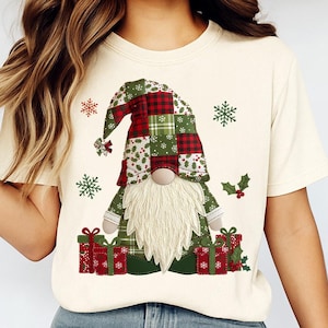 May include: Cream-colored t-shirt featuring a Christmas gnome design. The gnome has a patchwork hat and outfit in red, green, and white. Presents and snowflakes surround the gnome, creating a festive holiday theme.