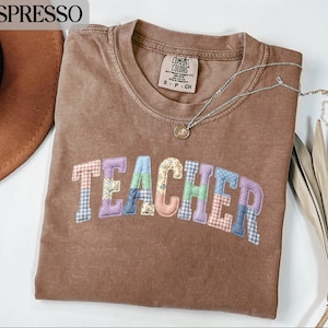 Może przedstawiać: Koszulka w kolorze espresso z napisem "TEACHER" w kolorowych literach patchworkowych. Koszulka ma okrągły dekolt i krótkie rękawy. Srebrny naszyjnik z małą zawieszką jest przerzucony przez koszulkę.