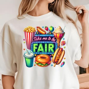 May include: White t-shirt with a colorful graphic design featuring fair food like popcorn, hot dogs, ice cream, and a donut. The graphic also includes a neon sign that says "Take me to the Fair".