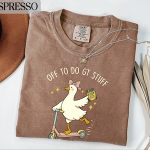 May include: An espresso-coloured t-shirt featuring a graphic of a white goose wearing a pink bow, riding a light blue scooter, and holding a box of crayons. The text above the goose reads OFF TO DO GT STUFF, accompanied by small yellow star icons.