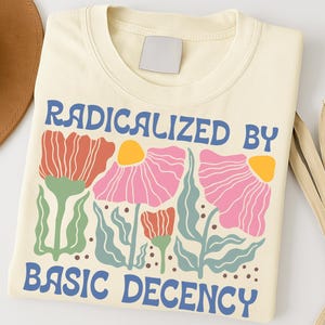 Può includere: T-shirt color crema con la scritta "RADICALIZED BY BASIC DECENCY" in blu. La maglietta presenta un design floreale con fiori rossi, rosa e gialli e foglie verdi. Un cappello marrone e una pianta secca sono sullo sfondo.