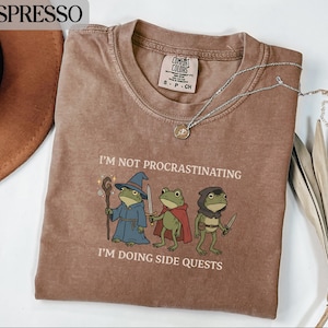 May include: Espresso-colored t-shirt with the text "I'M NOT PROCRASTINATING I'M DOING SIDE QUESTS" and a graphic of three frog characters. The frogs are dressed as a wizard, a knight, and a rogue. A silver necklace with a pendant is also visible.