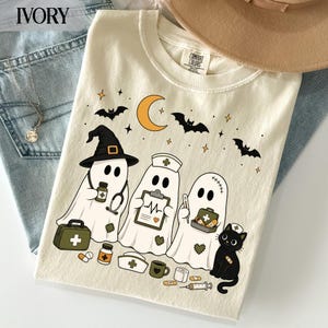May include: An ivory t-shirt featuring a graphic of three white ghosts dressed as medical professionals, including a doctor with a stethoscope, a nurse with a clipboard, and a ghost with a thermometer, alongside a black cat and medical supplies.