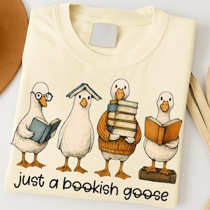 May include: A cream-coloured t-shirt with a cartoon illustration of four geese reading books. One goose wears glasses, another has a book on its head. The text "just a bookish goose" is printed below the geese in black.