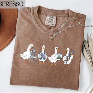 Puede incluir: Una camiseta marrón de cuello redondo con un gráfico de cuatro gansos blancos realizando actividades científicas. Un ganso usa un microscopio, otro sostiene un diagrama celular, un tercero lleva un cubo de cristal y el cuarto sostiene una cadena de ADN.