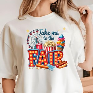 May include: White t-shirt with a colorful graphic design that says "Take me to the Fair". The graphic includes a Ferris wheel, popcorn, ice cream, and a hot dog.