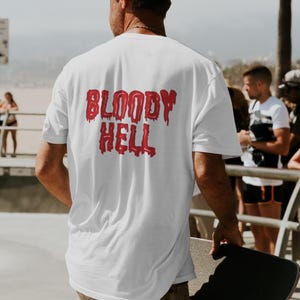 May include: White t-shirt with the red text "BLOODY HELL" in a dripping font. The shirt is worn by a person holding a skateboard. The person is wearing a beige cap and camouflage trousers.