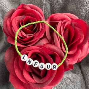 Peut inclure: Un bracelet en perles vertes avec des perles blanches qui épellent "LN FOUR". Le bracelet est sur une rose rouge.