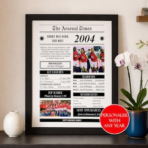 Op de afbeelding: Zwart-wit ingelijste print getiteld "The Arsenal Times" met het jaar 2004. De print bevat teamfoto's, belangrijke wedstrijddata en spelertransfers. Een rode cirkel met tekst staat in de rechter benedenhoek.