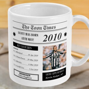 May include: A white ceramic mug with a black and white newspaper design. The mug features a headline that reads "The Toon Times" and a subheading that reads "SCOTT WAS BORN 15TH MAY! 2010". The mug also includes a photo of a soccer player celebrating a goal.