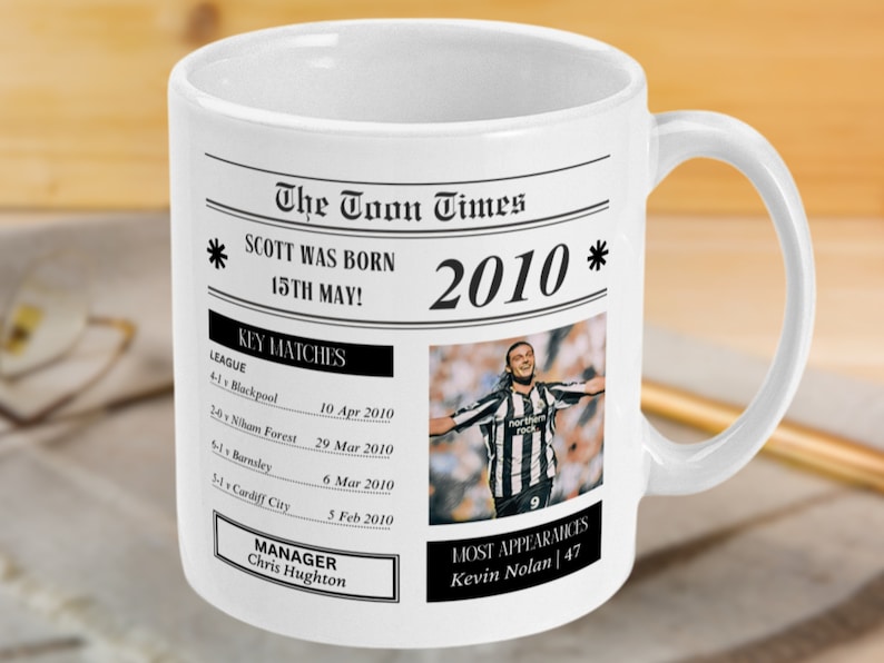 May include: A white ceramic mug with a black and white newspaper design. The mug features a headline that reads "The Toon Times" and a subheading that reads "SCOTT WAS BORN 15TH MAY! 2010". The mug also includes a photo of a soccer player celebrating a goal.