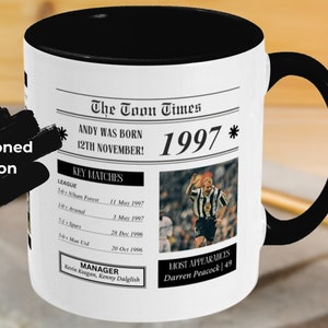 May include: A black and white mug with a newspaper design featuring the text "The Toon Times" and "1997". The mug has a black handle and a black rim.