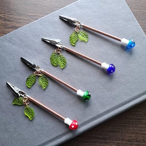 May include: Four decorative alligator clips with copper-colored flexible stems. Each clip has a different colored mushroom-shaped end: red, green, blue, and blue. Each clip also has a charm of three green glass leaves.