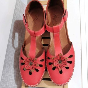 Könnte beinhalten: Rote Ledersandalen mit floralem Design und Aussparungen. Die Sandalen haben einen T-Riemen, einen verstellbaren Knöchelriemen und eine dekorative Blume. Die Sohlen sind braun und die Nähte sind weiß.