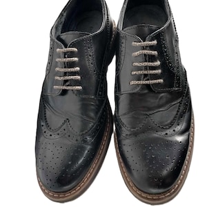 Peut inclure: Une paire de chaussures richelieu en cuir noir avec des perforations décoratives de type brogue sur le bout et les côtés, un bout arrondi et des lacets texturés marron clair. Les chaussures ont un petit talon et une trépointe marron visible.