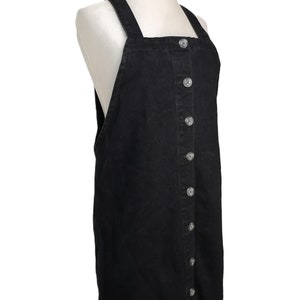 Op de afbeelding: Zwarte denim overalljurk met knoopsluiting aan de voorkant en verstelbare bandjes.