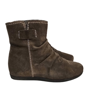 May include: Brown suede ankle boots with a low heel. The boots feature a ruched design on the upper and a small strap detail with a buckle. White stitching accents the seams and the top of the boot.