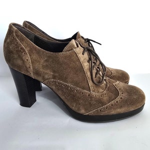 May include: Brown suede oxford-style heeled shoes with a lace-up design. The shoes feature a rounded toe, a stacked heel, and decorative perforations along the edges. The heels are approximately 3 inches high.