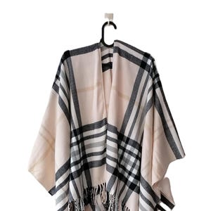 Puede incluir: Un poncho beige con un estampado de cuadros negros, grises y blancos. El poncho tiene un dobladillo con flecos y está colgado de una percha negra. La prenda está hecha de un material suave.