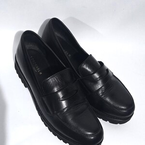 May include: A pair of black leather loafers with a rounded toe and a slip-on design. The shoes have a textured rubber sole and the word "STOCKHOLM" is visible on the side. The shoes are set against a white background.