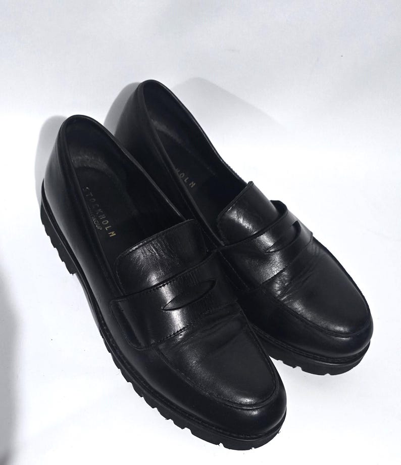 May include: A pair of black leather loafers with a rounded toe and a slip-on design. The shoes have a textured rubber sole and the word "STOCKHOLM" is visible on the side. The shoes are set against a white background.
