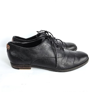 Puede incluir: Un par de zapatos Oxford de cuero negro con cierre de cordones. Los zapatos tienen una puntera redondeada y un tacón bajo. El talón presenta un pequeño detalle de cuero marrón. Los zapatos están sobre un fondo blanco.