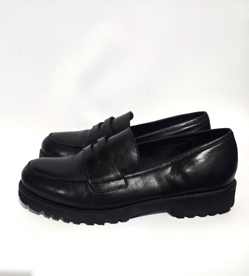 May include: A pair of black leather penny loafers. The shoes have a classic design with a rounded toe, a strap across the vamp, and a chunky, lugged sole. The leather appears smooth and polished.
