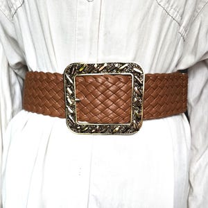 May include: A wide brown braided belt featuring a large, rectangular gold-tone buckle. The buckle has a textured, metallic border with a dark, shimmering inlay. The belt strap is constructed from a woven, interlaced material in a solid brown color.