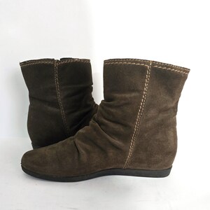 May include: A pair of brown suede ankle boots. The boots have a rounded toe, a low heel, and a slouchy design. The stitching is visible in a lighter color, and the boots are set against a white background.