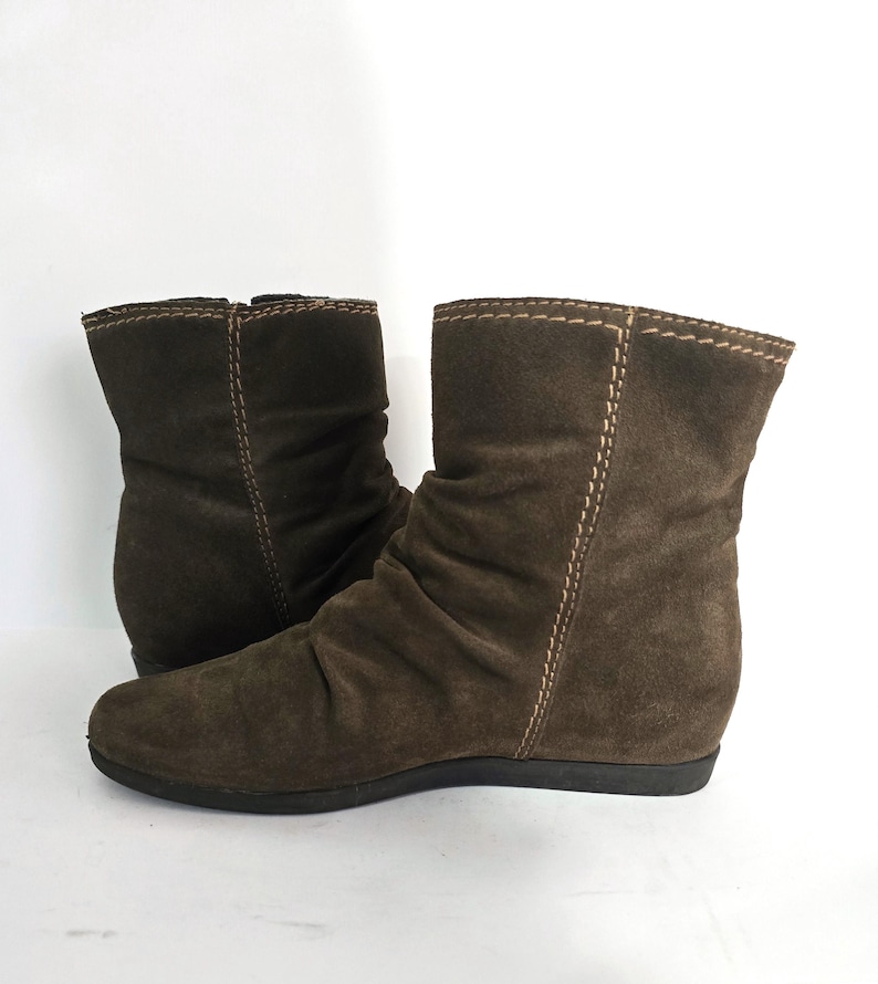 May include: A pair of brown suede ankle boots. The boots have a rounded toe, a low heel, and a slouchy design. The stitching is visible in a lighter color, and the boots are set against a white background.