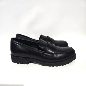 May include: A pair of black leather loafers with a rounded toe and a thick, lugged sole. The shoes have a classic penny loafer design with a strap across the vamp. The shoes are set against a white background.