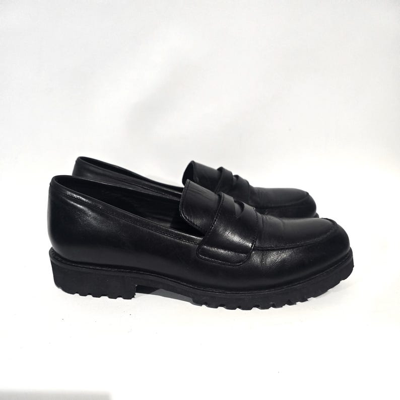 May include: A pair of black leather loafers with a rounded toe and a thick, lugged sole. The shoes have a classic penny loafer design with a strap across the vamp. The shoes are set against a white background.
