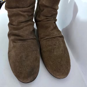 May include: A pair of brown suede boots. The boots have a slouchy design with a rounded toe and a mid-calf height. The suede has a soft, textured appearance, and the boots appear unworn.