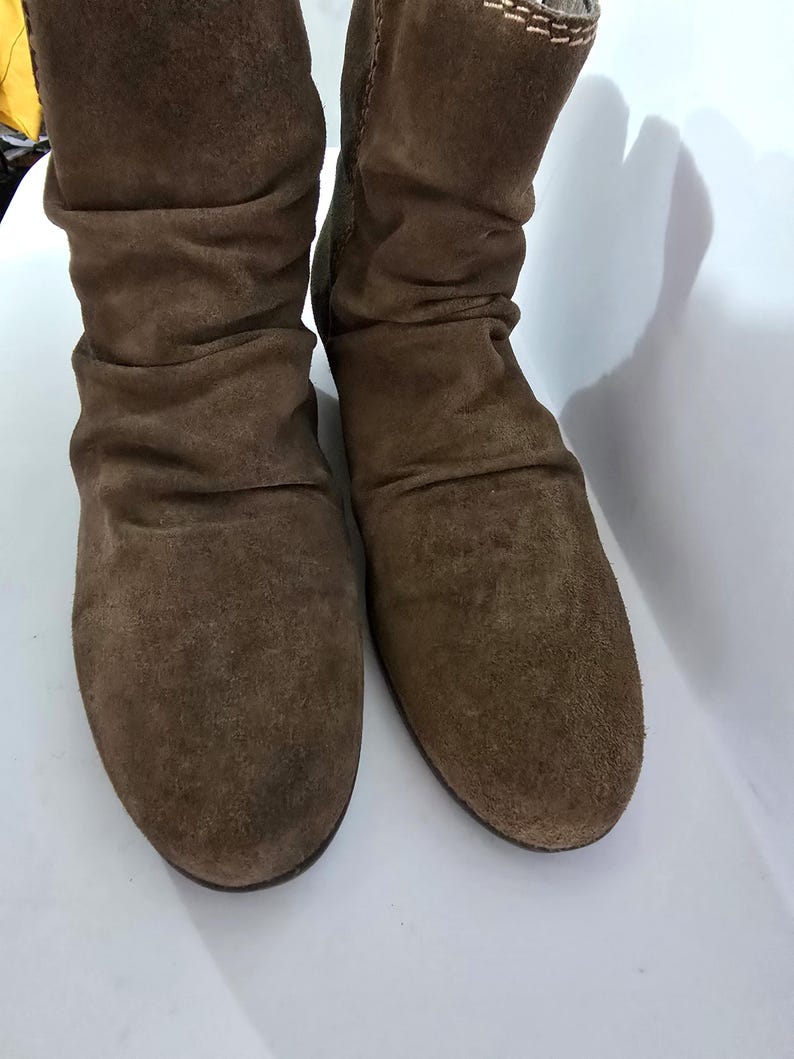 May include: A pair of brown suede boots. The boots have a slouchy design with a rounded toe and a mid-calf height. The suede has a soft, textured appearance, and the boots appear unworn.