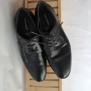 May include: A pair of black leather lace-up shoes with the brand name "Kingsmen" printed on the inside of each shoe.