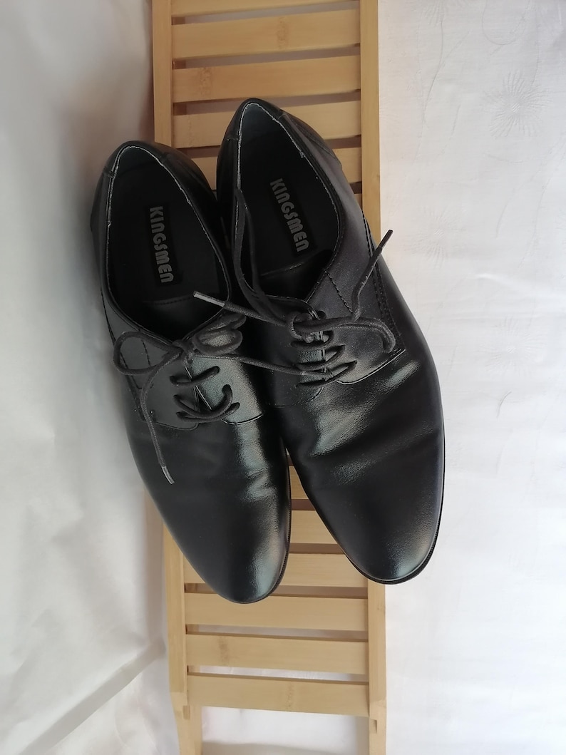 May include: A pair of black leather lace-up shoes with the brand name "Kingsmen" printed on the inside of each shoe.