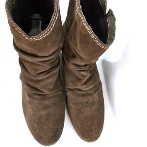 May include: A pair of brown suede boots with a slouchy design. The boots have a mid-calf height and a rounded toe. The edges are stitched with a contrasting thread. The boots are set against a white background.