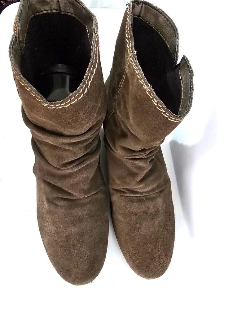 May include: A pair of brown suede boots with a slouchy design. The boots have a mid-calf height and a rounded toe. The edges are stitched with a contrasting thread. The boots are set against a white background.