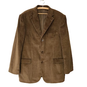 Könnte beinhalten: Brauner Cord-Blazer mit Zwei-Knopf-Verschluss, zwei Vordertaschen und einem fallenden Revers. Die Jacke hängt an einem Holzbügel vor weißem Hintergrund. Der Stoff des Blazers hat eine vertikale Rippenstruktur.